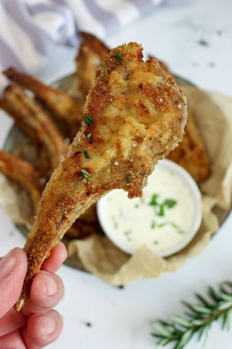 A crumbed lamb cutlet is held in front of more lamb cutlets are served in a bowl with a bowl of dipping sauce