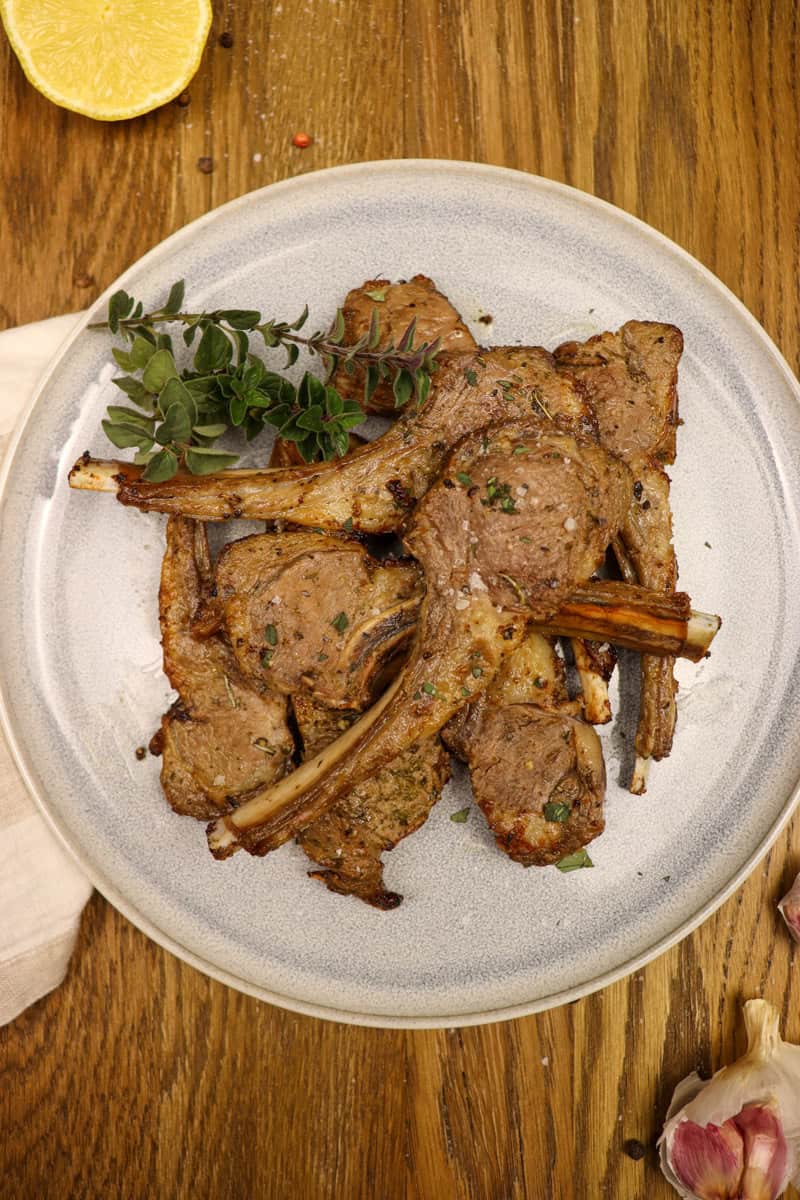 The cooked lamb cutlets are served on a plate alongside fresh oregano. There is some fresh garlic and lemon on the table to the side of the plate