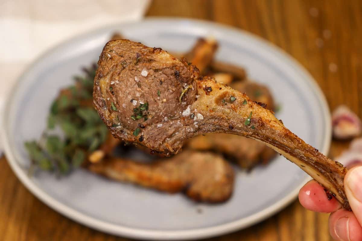 A cooked lamb cutlet is held close to the camera with the dish in the background