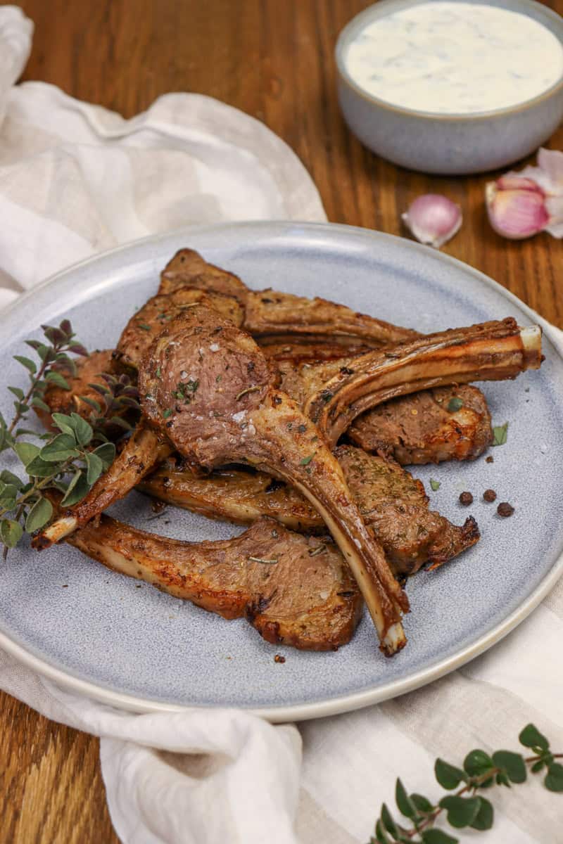 The cooked lamb cutlets are served on a plate alongside fresh oregano. There is a bowl of dipping sauce and some garlic in the background