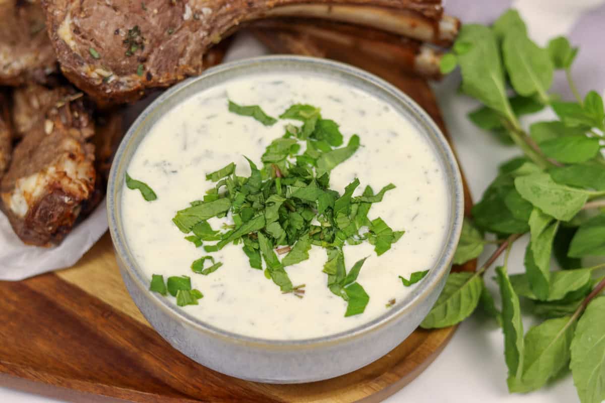A bowl of mint and yogurt sauce served next to some lamb cutlets with fresh mint on the side