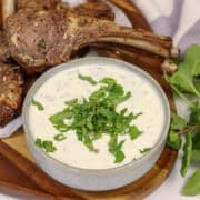 A bowl of mint and yogurt sauce served next to some lamb cutlets with fresh mint on the side