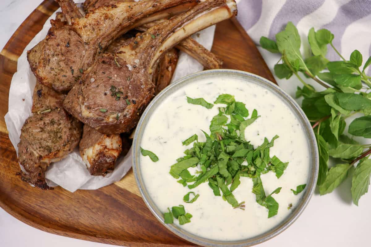 A bowl of mint and yogurt sauce served next to some lamb cutlets with fresh mint on the side