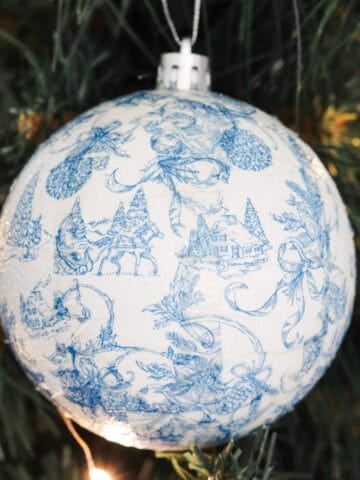 A DIY paper napkin decoupage Christmas tree ornament in blue and white with Christmas motifs hanging in a Christmas tree